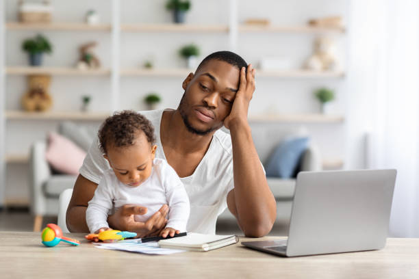 Tired african american father with infant baby working on laptop at home office, exhausted black freelancer man sitting at desk with computer and holding little child, having multitasking stress